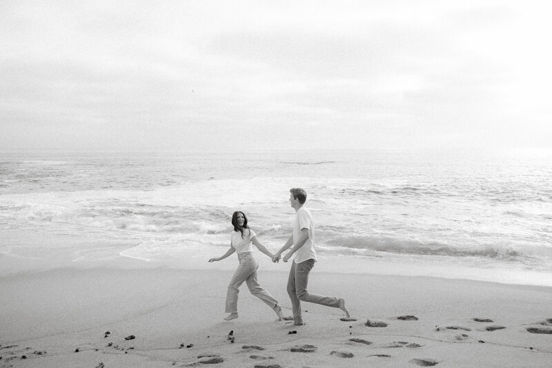 Black and white photo of man and woman holding hands running on the beach