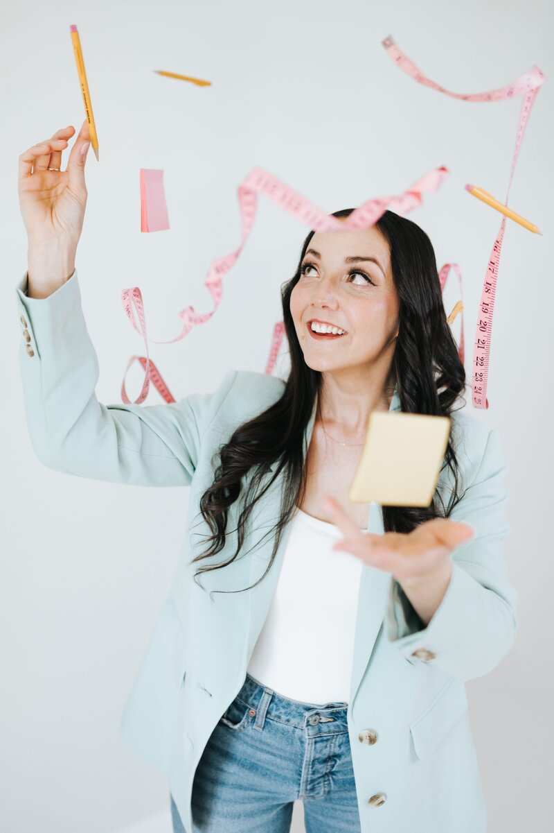Woman in pastel blue blazer smiles up while reaching for pencil as several other pencils, post its and measuring tapes are suspended above her.