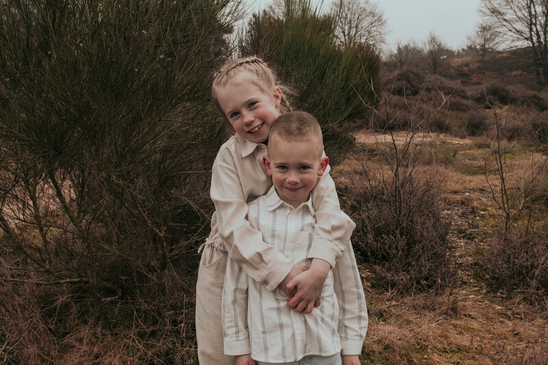 Twee kinderen staan in een veld, omringd door gras en bloemen, met een heldere lucht op de achtergrond.