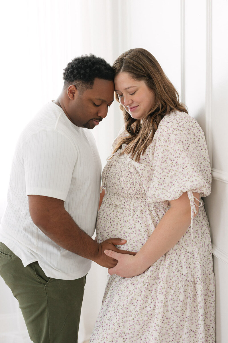 Mom and dad embracing mom's pregnant belly as she leans against a wall in a bright louisville photography studio