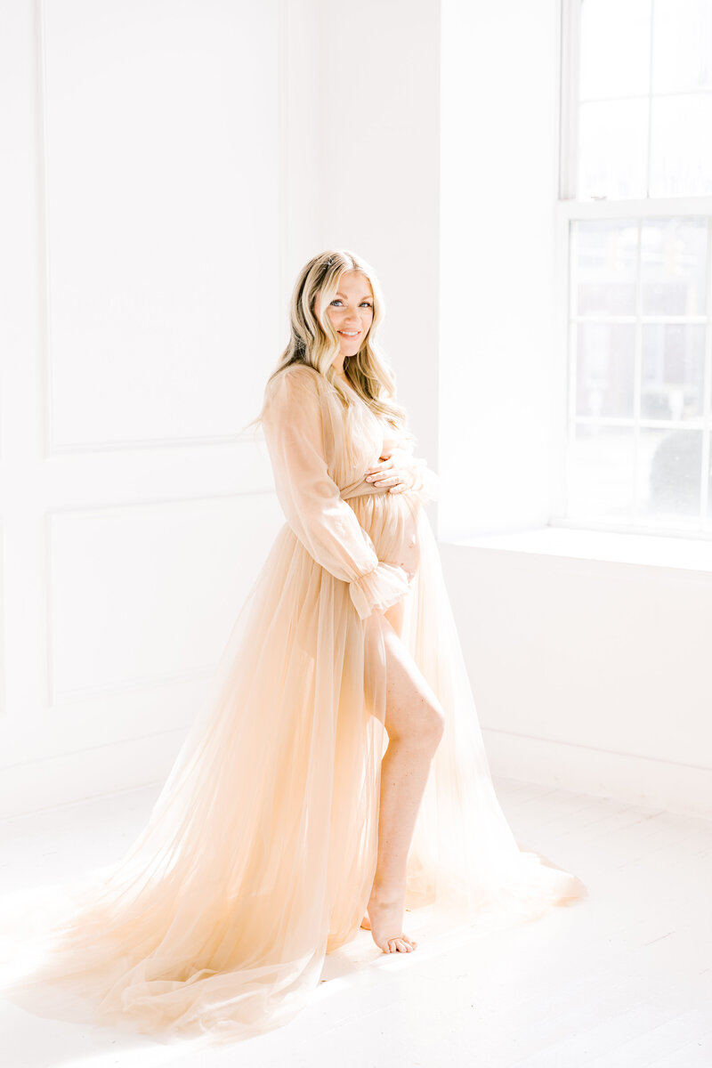 A Charlotte Maternity Photographer image of a woman with a popped leg standing in a window smiling in a pink maternity gown