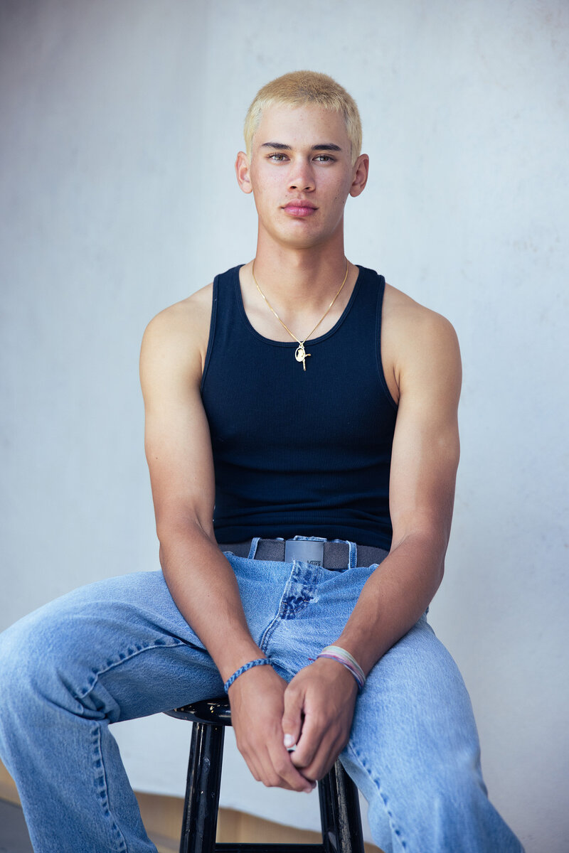 Teen guy in a black tank top and light blue jeans sitting casually against a light gray wall, wearing a cross necklace and wristbands.