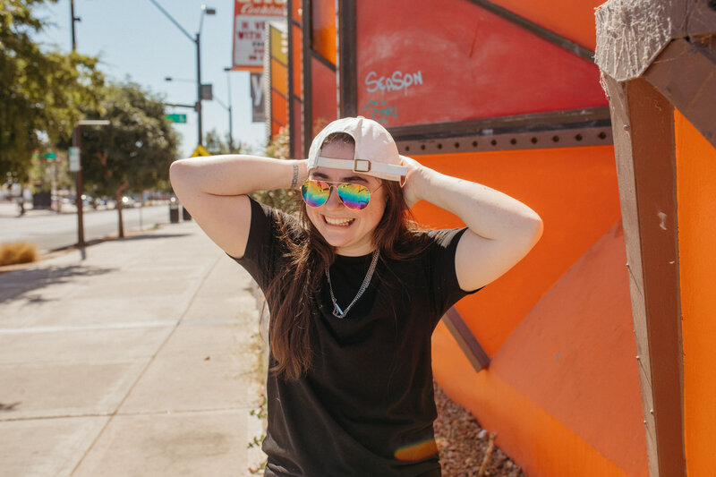 Girl smiling with rainbow sunglasses in front of a red building.