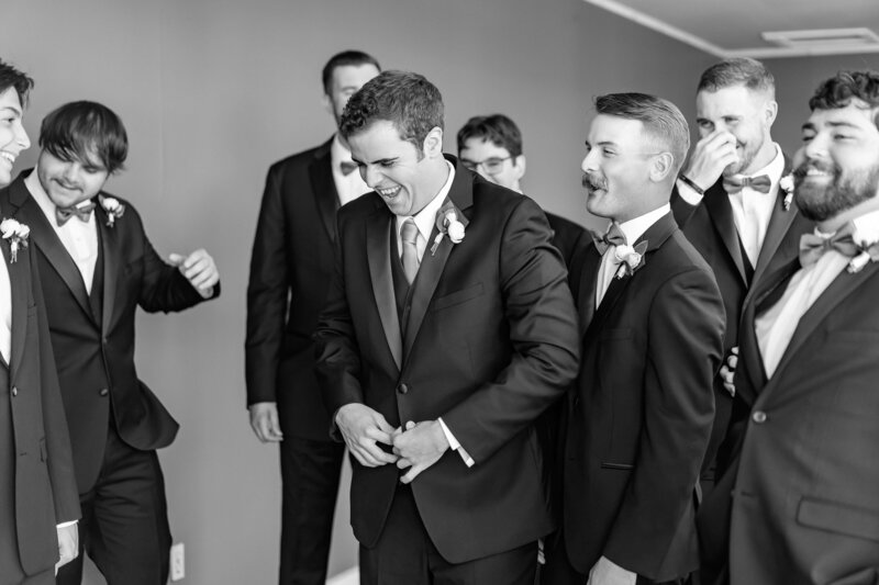 A group of men in suits and boutonnieres stand together, laughing and smiling. The atmosphere appears joyful and celebratory. The photo is in black and white.