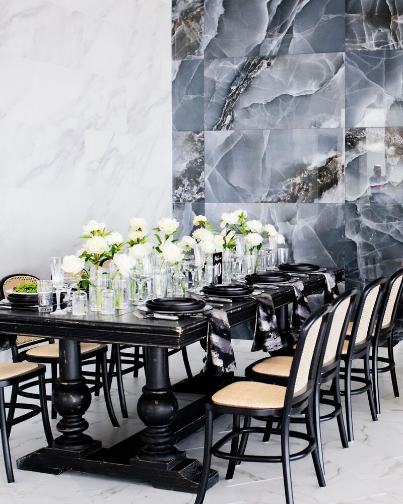 Black rattan-back chairs around black wood tables styled with white florals and black plates against a marble wall backdrop.