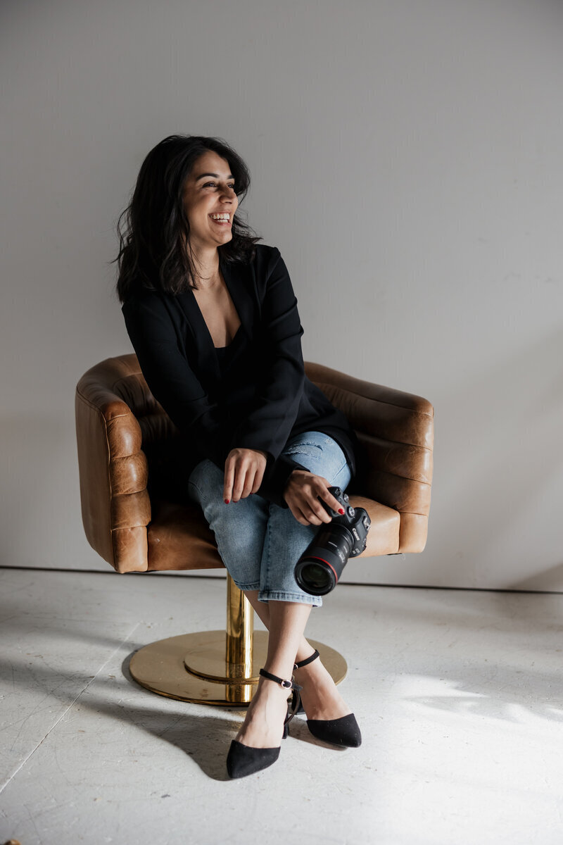 A person with long dark hair, embodying the New England lifestyle photographer vibe, captures a couple sitting closely in a cozy, rustic bar. A newspaper rests on a wooden table in the foreground, while an exit sign and ceiling fan hint at the room's characterful charm.