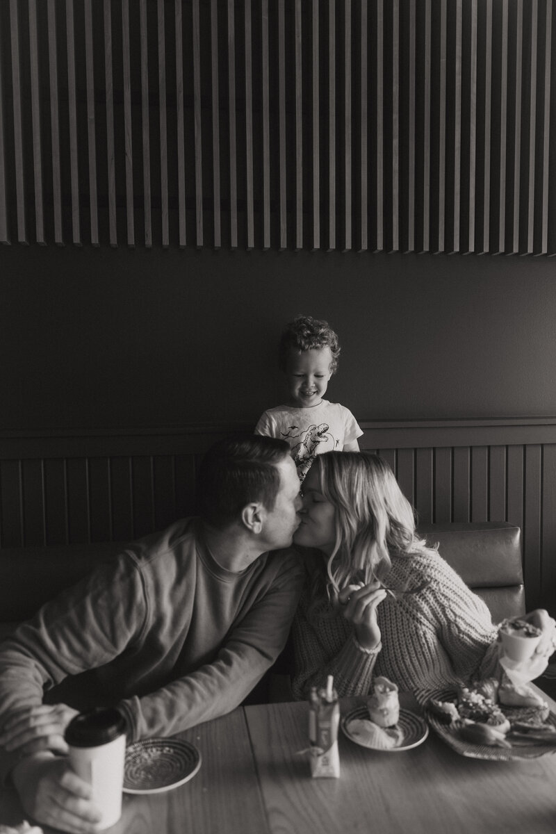 Couple kisses in a diner while young boy smiles from behind them.