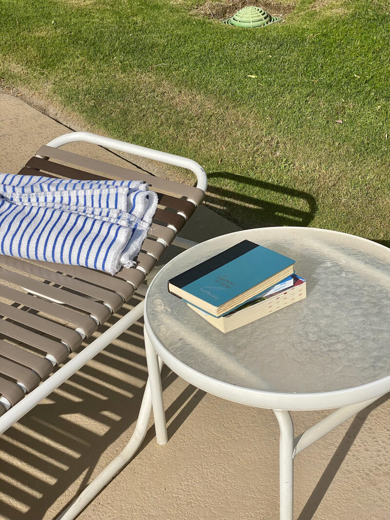Lounge chair with a striped towel beside a small table holding two books, placed outdoors on a sunny day