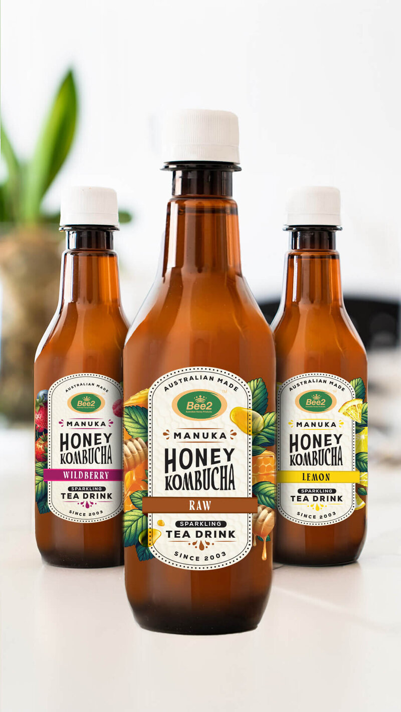 Three bottles of Manuka Honey Kombucha are displayed in a bright, minimalistic setting. The bottles showcase different flavors, including the original, Lemon, and Wildberry, emphasizing the variety and organic nature of the kombucha drinks.