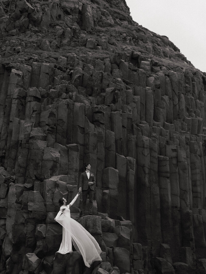 Bride and groom standing on towering basalt rock columns in a dramatic black-and-white elopement portrait, with the bride’s dress flowing in the wind.