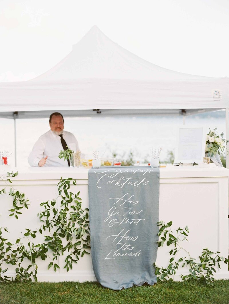 custom white wedding bar at lakefront reception blue linen with white calligraphy