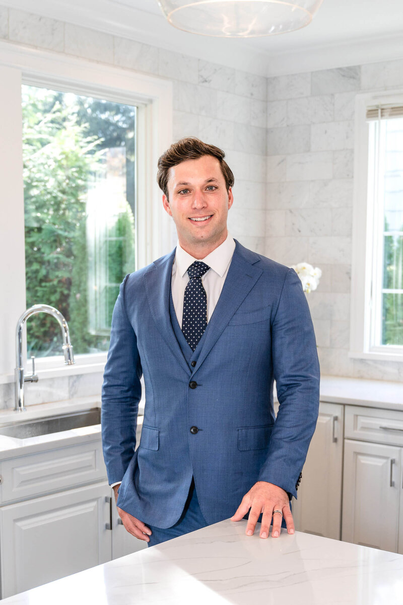 A young professional man in a  blue suit, standing at the edge of a marble white countertop,  in an all white kitchen smiling while posing for Branding photos in Greenwich, CT.