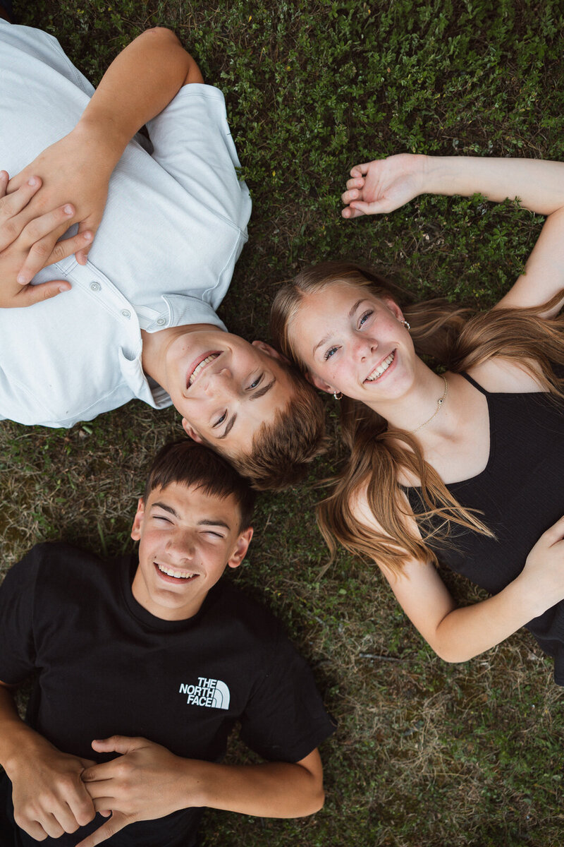 Three siblings laying in grass looking up at the camera smiling. 