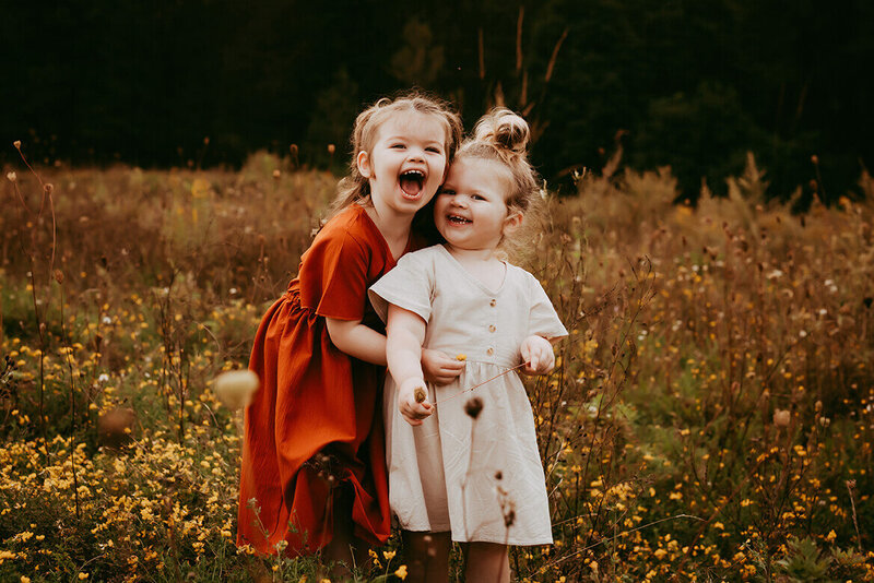 A portrait of two sisters standing in yellow wild flowers hugging and smiling