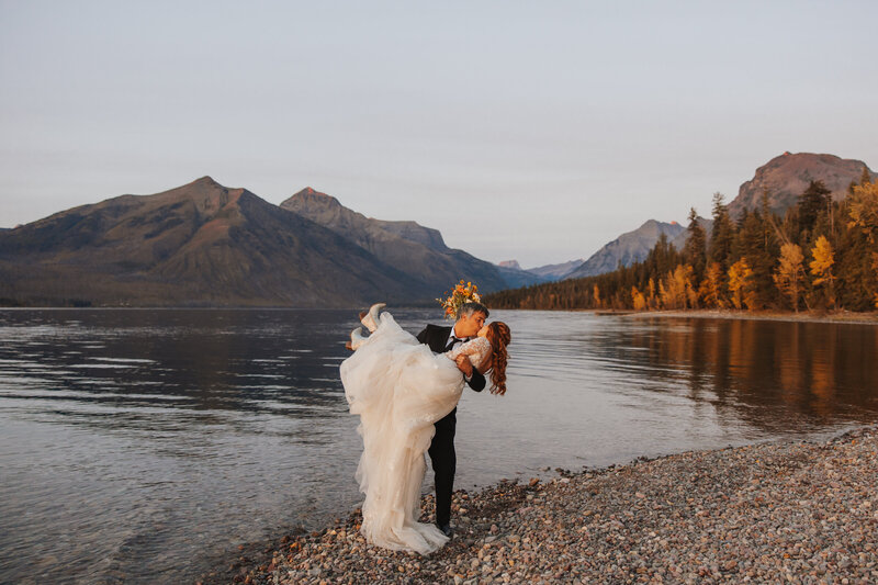 Couple on a Montana lakeshore as the groom holds and kisses the bride at sunset.