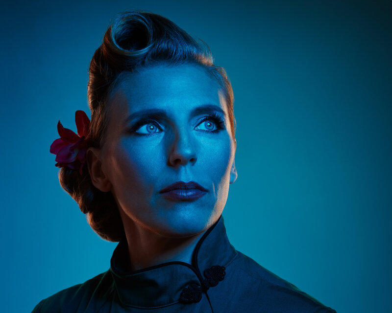 Dramatic portrait of Ylva Erevall with moody blue lighting, wearing a vintage-inspired hairstyle and looking thoughtfully into the distance.