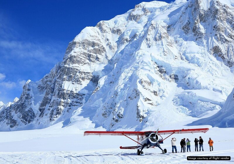 Images of a red and white ski plane landed on the snowfield glacier in Denali National Park.  There are people standing under the right wing of the plane--they look tiny next to the plane, with huge, snowcovered craggy mountains shooting up behind them out of the glacier.  The mountains fill nearly the entire back of the frame, except for a tiny peek of bluebird blue sky between the Alaska mountain peaks, a scene perfect for a non-helicopter glacier elopement in Alaska. 