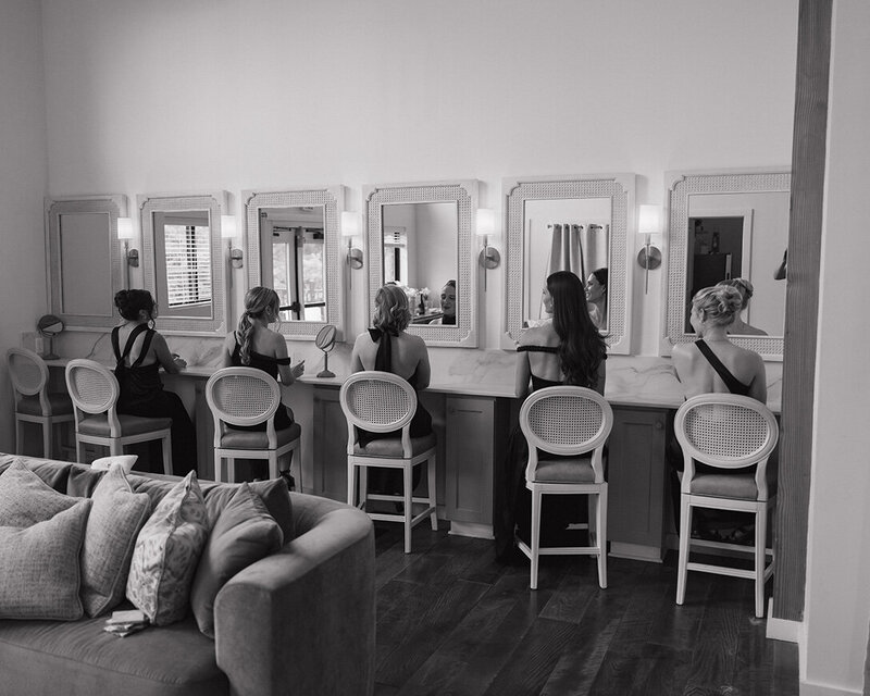 Five women in bridesmaid dresses sit at a vanity with mirrors, preparing for a Columbus wedding. The room has an elegant and serene atmosphere.