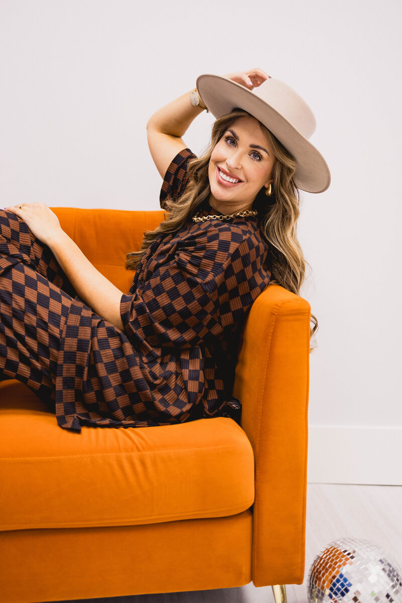woman wearing a wide brimmed hat sitting in an orange chair while smiling