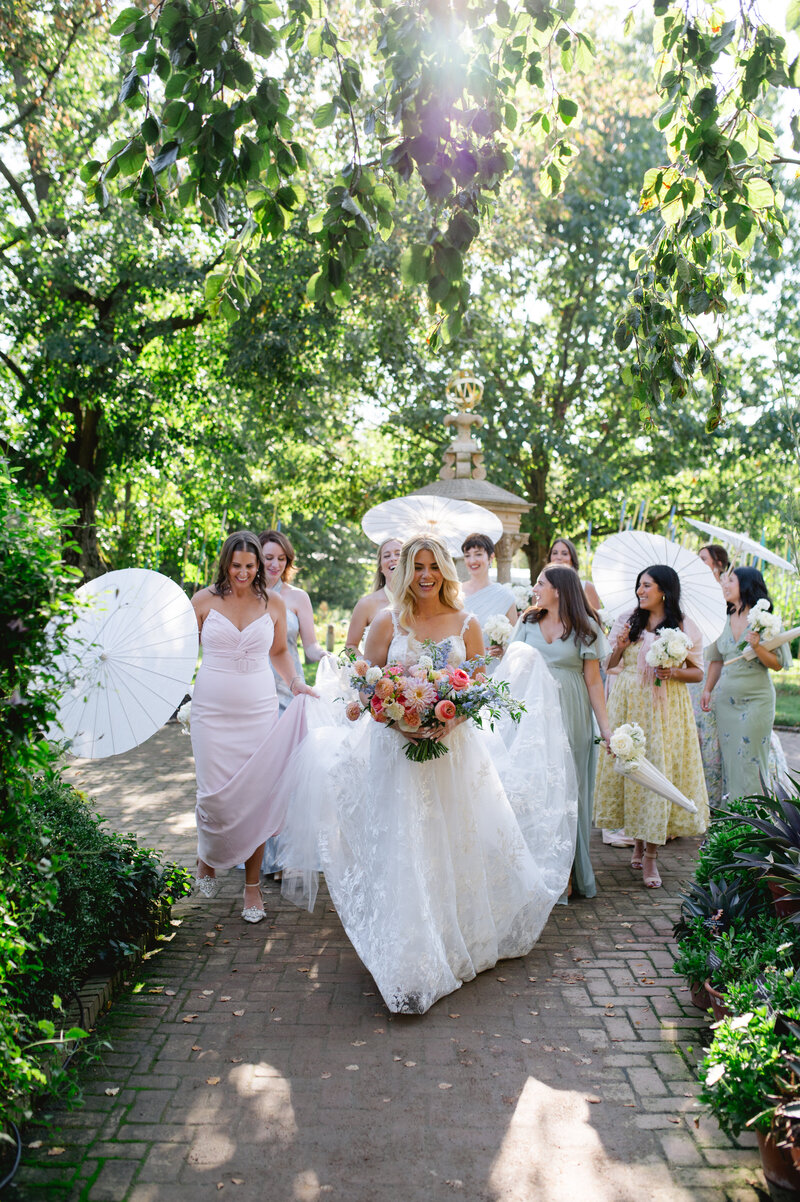 A bride holding flowers walking in a park setting with women walking behind her holding her dress. 