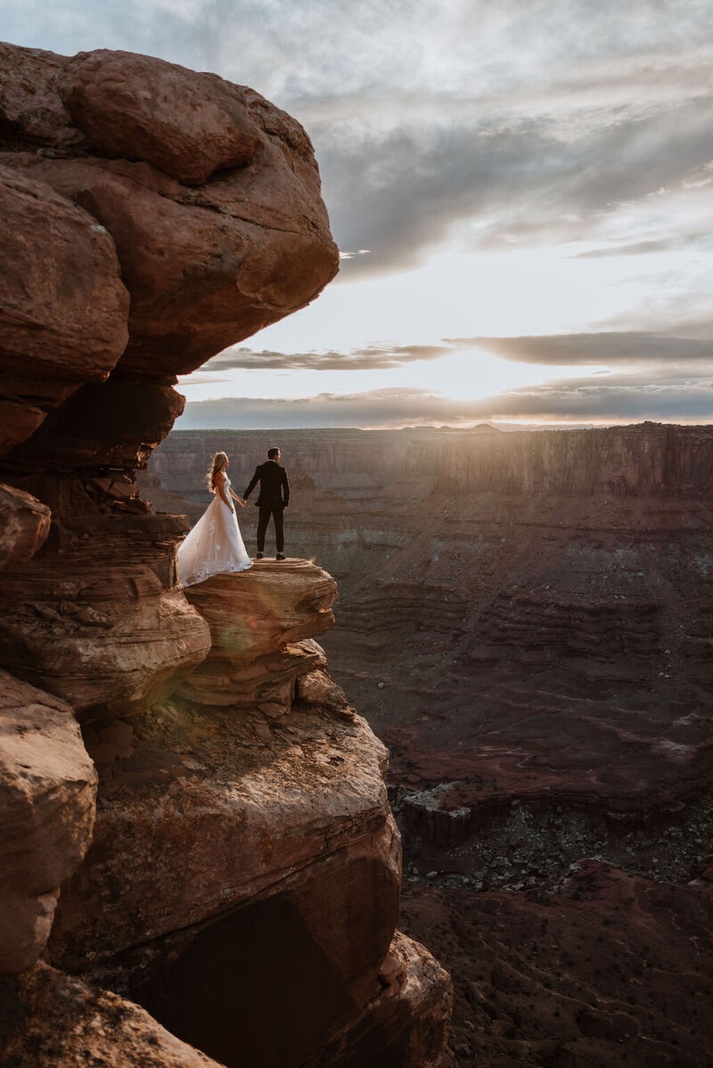 bride and groom standing on large rock formation cliff with the desert stretch out before them