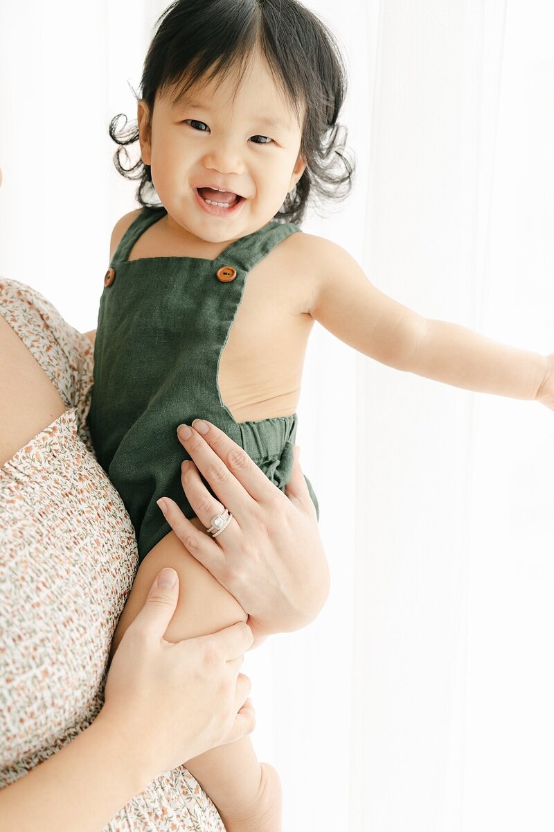 One year old baby boy in green overalls smiles at Indianapolis family photographer, Brittney Lear, while being held by his mom.