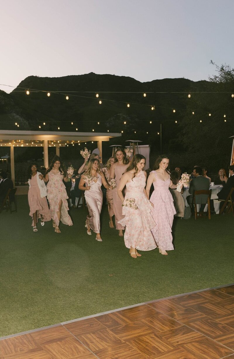 Bridesmaids running to the outdoor dance floor in pink with pink flowers at night