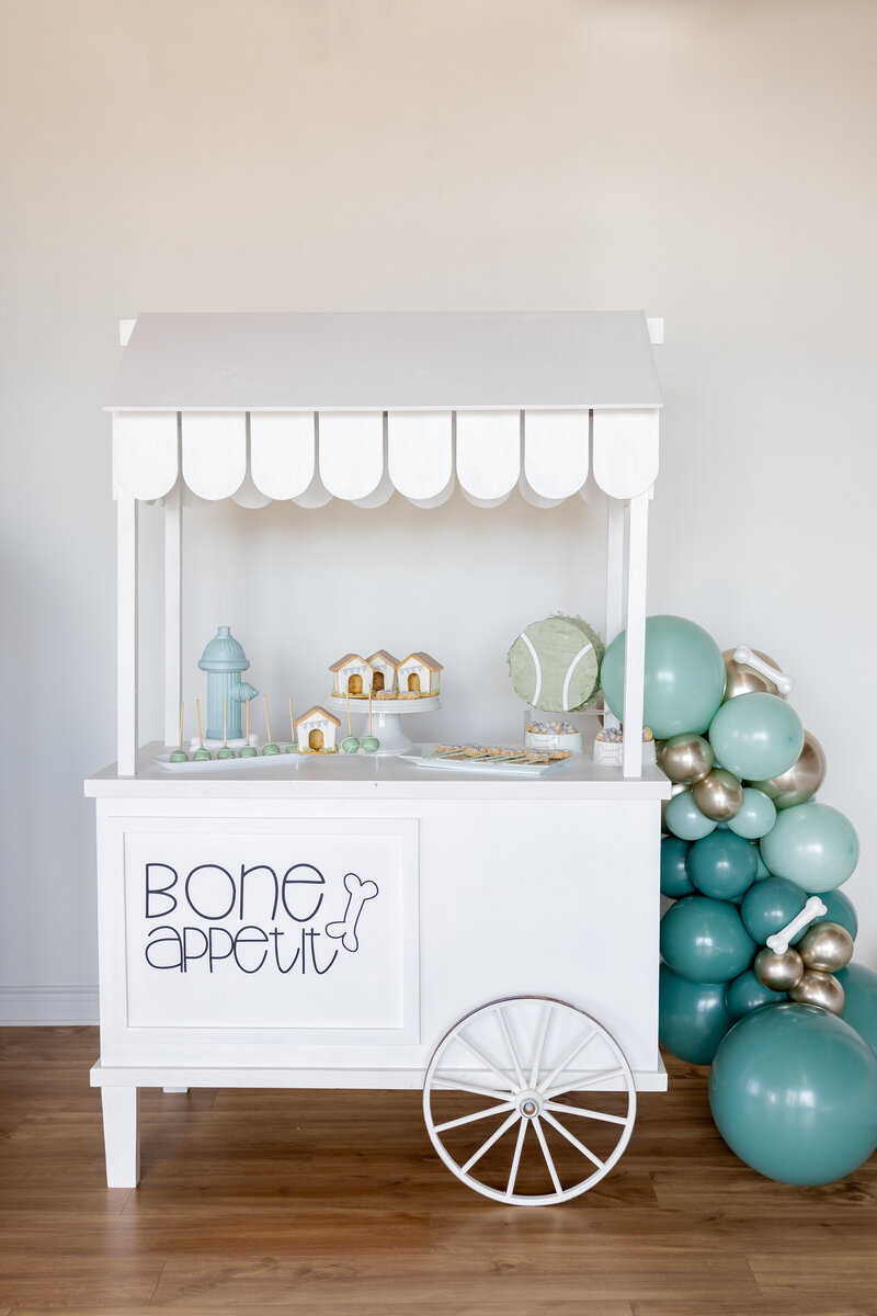 White dessert cart with cookies, rice krispie treats, cake balls, tennis ball pinata, dog decorations and blue balloons,
