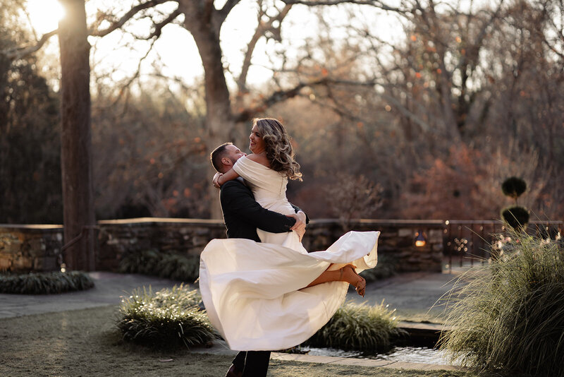 groom holding bride up and spinning her around. Her dress is twirling in the wind with the sunset in the back of them.