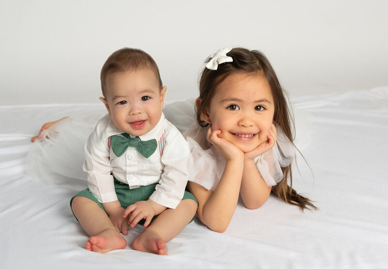 Indoor asian siblings created by Ballantyne childrens photographer Insley Photo boy in green bow tie just sitting up and girl in white tulle dress, bow and smiling with chin in her hands next to her brother.