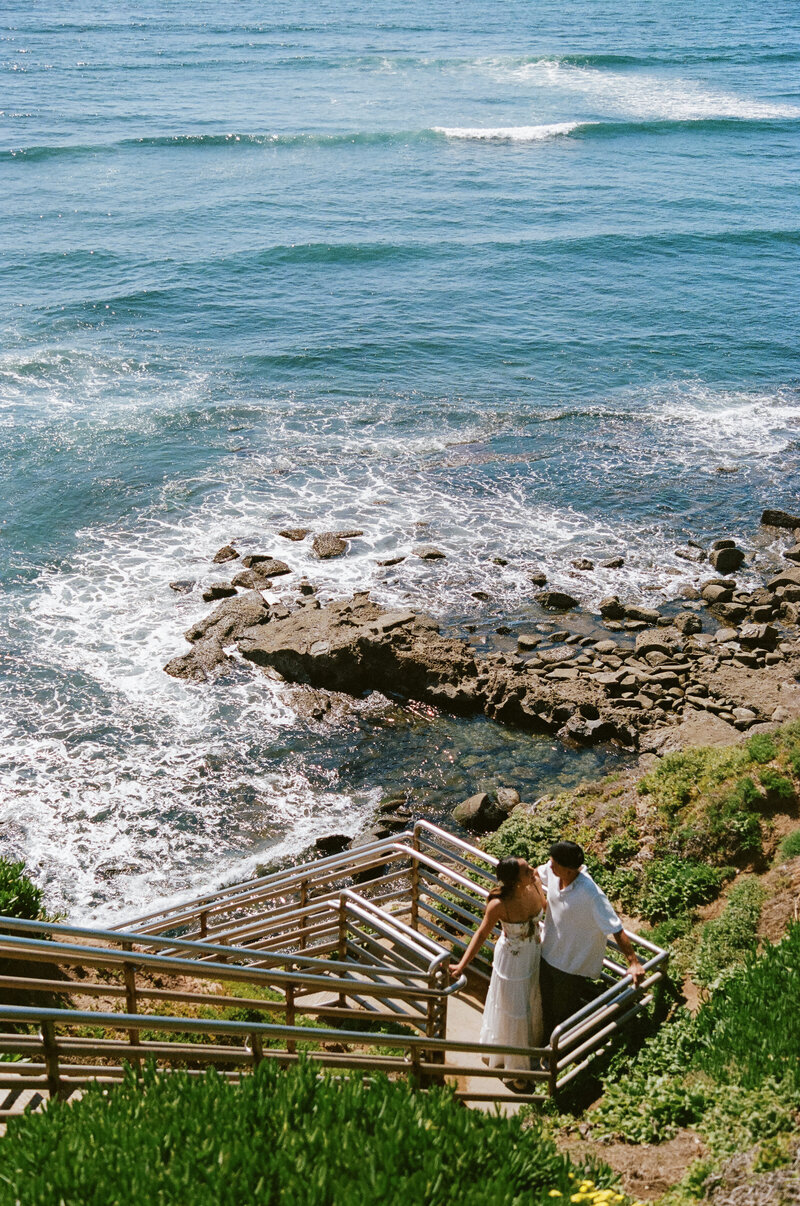 A 35mm film photo of a couple descending a staircase to a rocky beach in San Diego, with the ocean waves hitting the shore, capturing a serene and intimate moment during their engagement session.