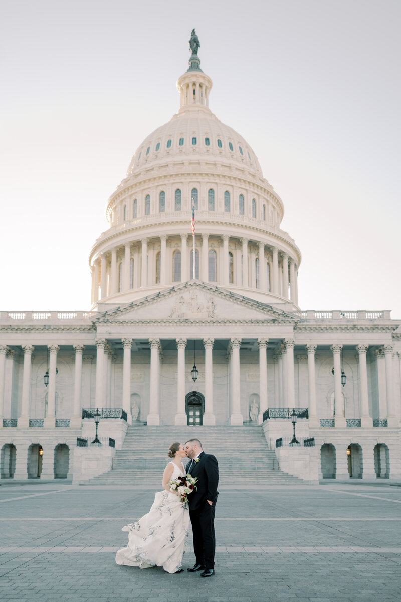 bride and groom kissing in front of the capitol in washington dc. the bride is wearing a unique dress with an elegant print on the skirt and beautiful v neck front. The groom is wearing a black tux and black shoes. The bride is holding a bouquet of white and deep burgundy florals perfect for their winter wedding. The photo was taken by washington dc wedding photographer omar & company