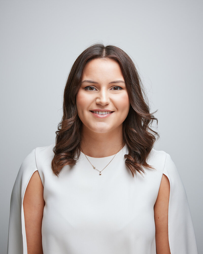 The image is a portrait of a smiling lady against a wall. She is wearing a blouse and a necklace.