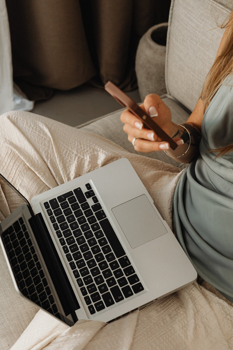 woman in linen pants on a couch with a laptop, looking at her mobile phone