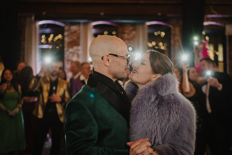 A newlywed couple shares a kiss after a Jewish wedding at the Boathouse at Confluence Park surrounded by guests holding sparklers at night, creating a magical and festive atmosphere. The bride's curly hair and the groom's beard are softly illuminated by the warm light of the sparklers.