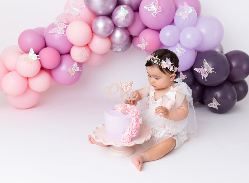 Rochel Konik Photography | Top Brooklyn Cake Smash Photographer captures baby girl in white lace romper and floral crown getting messy eating her first birthday cake in a studio cake smash session. Baby has icing on her hands , feet, and legs and is looking up at the  cake. Pink, lavender balloon garland