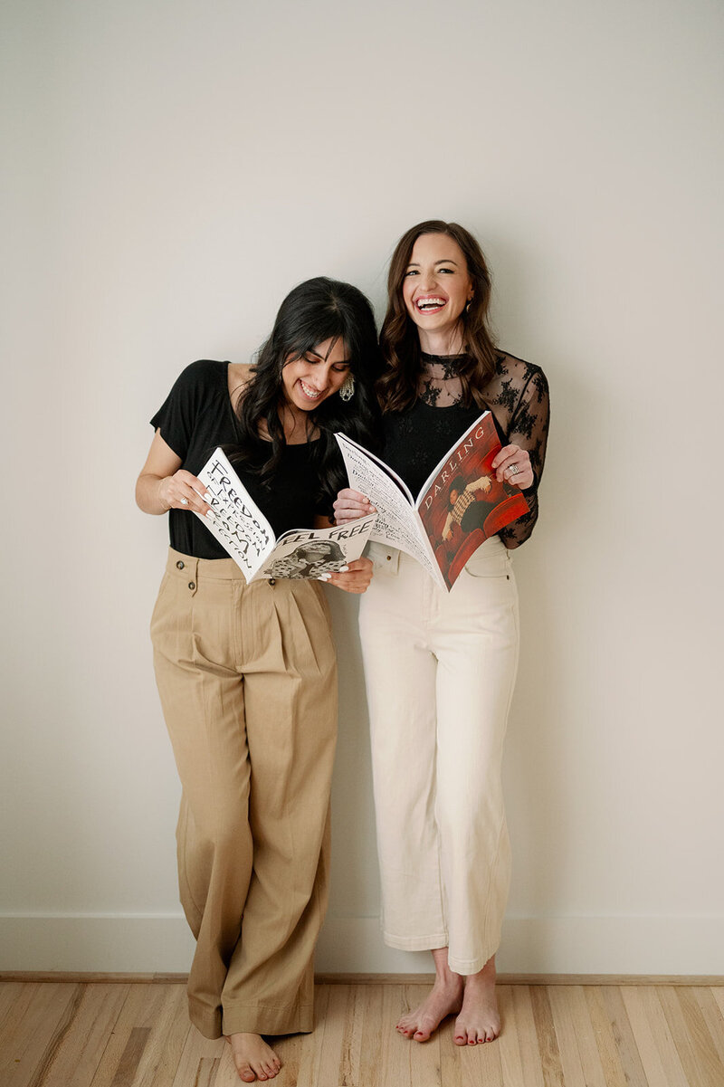 Two women lean against a wall and look at magazines