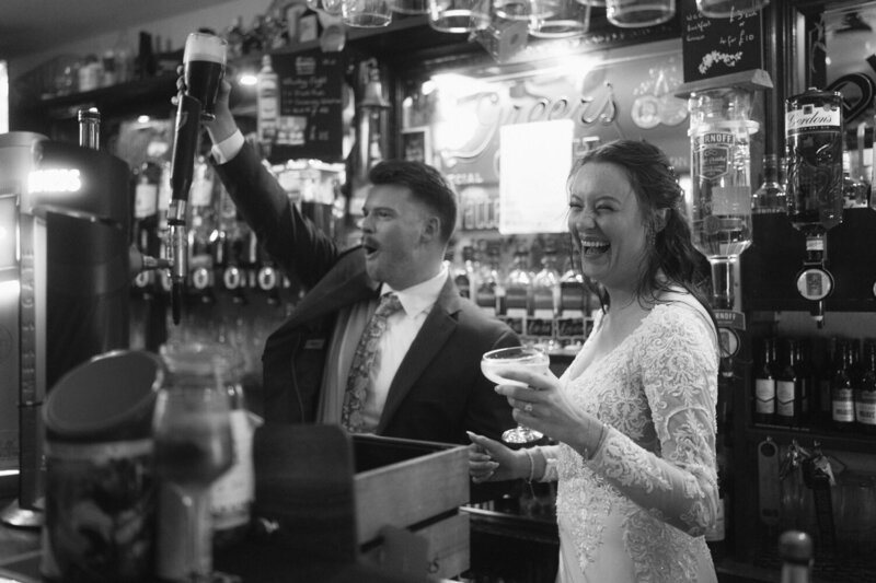 Unique Elopement Ideas | Bride and groom laughing and standing behind the bar in a pub in Ireland as groom holds up a pint of Guinness beer he just drank
