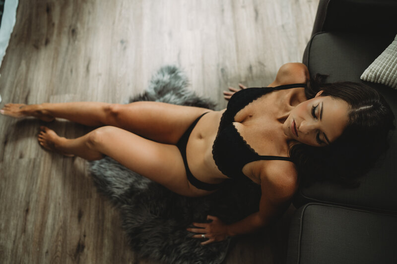 A woman in black lingerie sits on a rug while leaning back onto a leather chair
