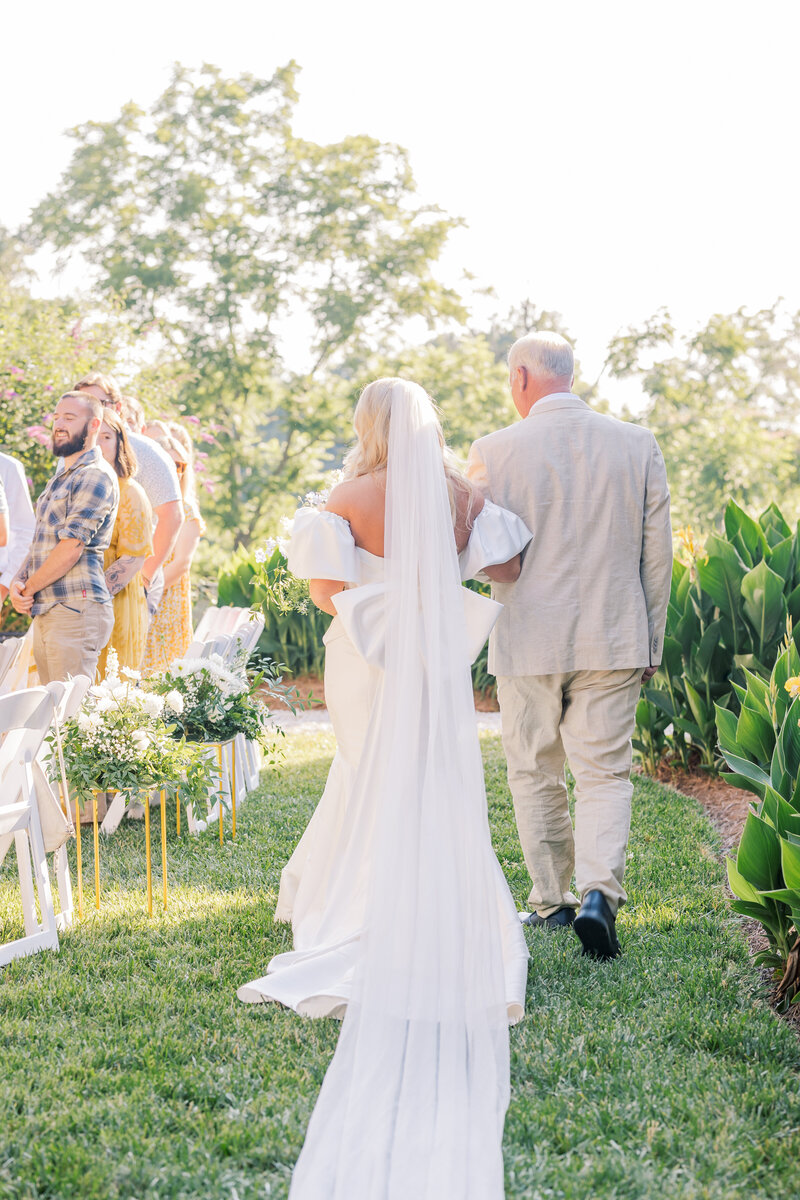 A bride walks down the aisle with her father in an outdoor ceremony, her long veil and elegant gown flowing behind her as guests look on under the golden sunlight.