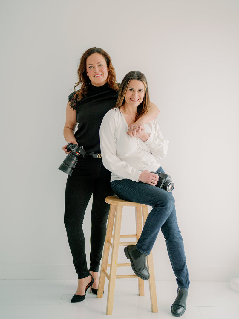 Photography team photo with one sitting and the other leaning elbow on her shoulder while smiling at camera