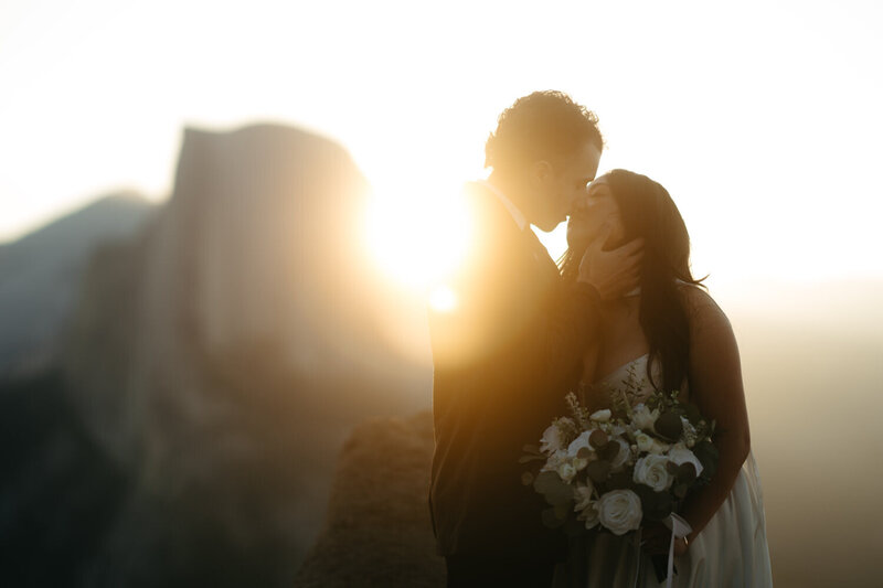 Close up photo of a Yosemite elopement, groom is holding brides face to kiss her with a sun flair and Half Dome behind them