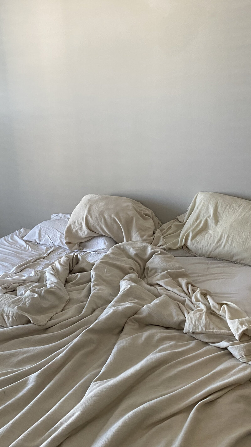 Unmade bed with crumpled beige sheets and pillows against a plain, light-colored wall. The scene conveys a sense of calm disorder and morning light.