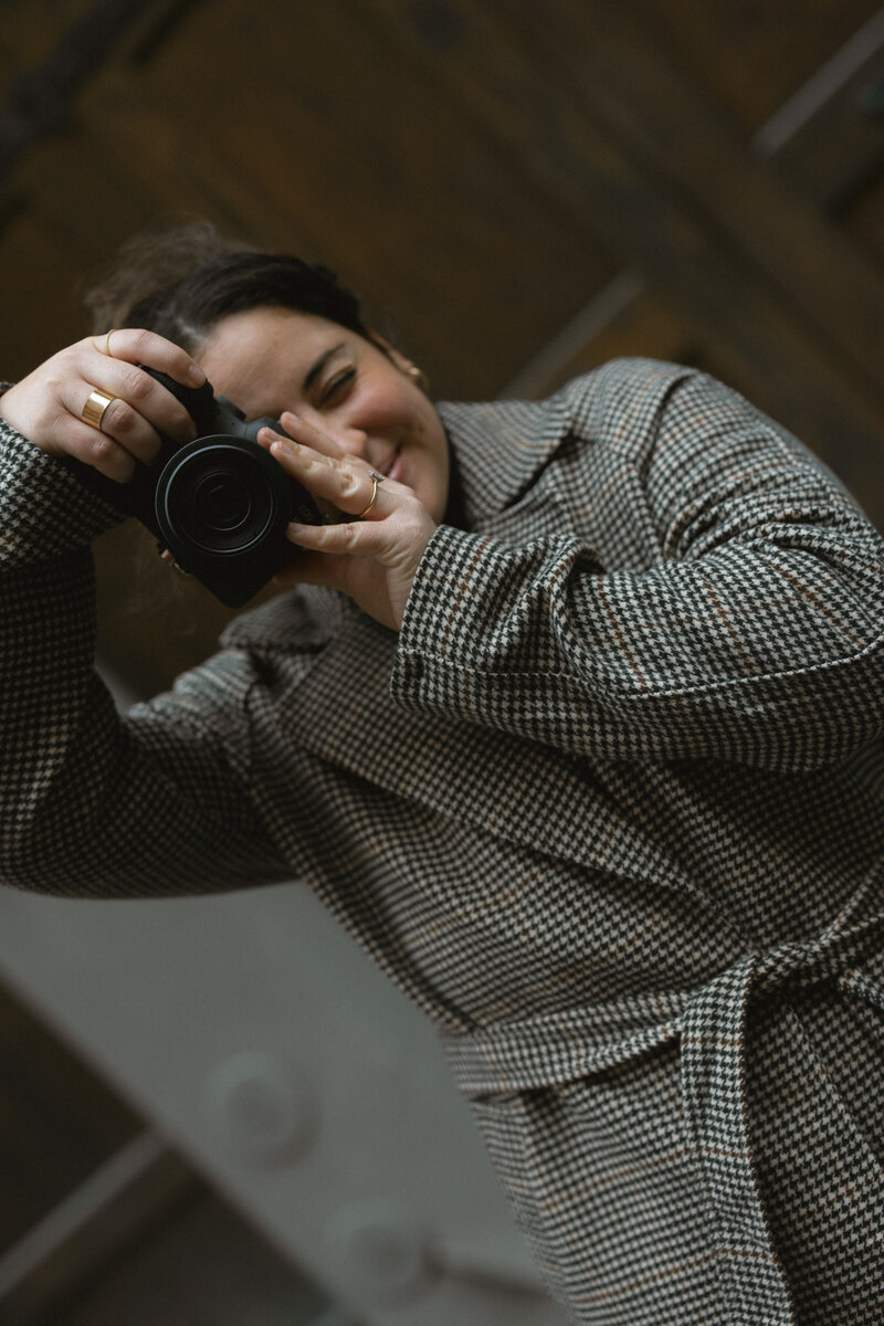 A photographer wearing a stylish checkered coat holds a camera, capturing an image in a cozy, wooden interior.