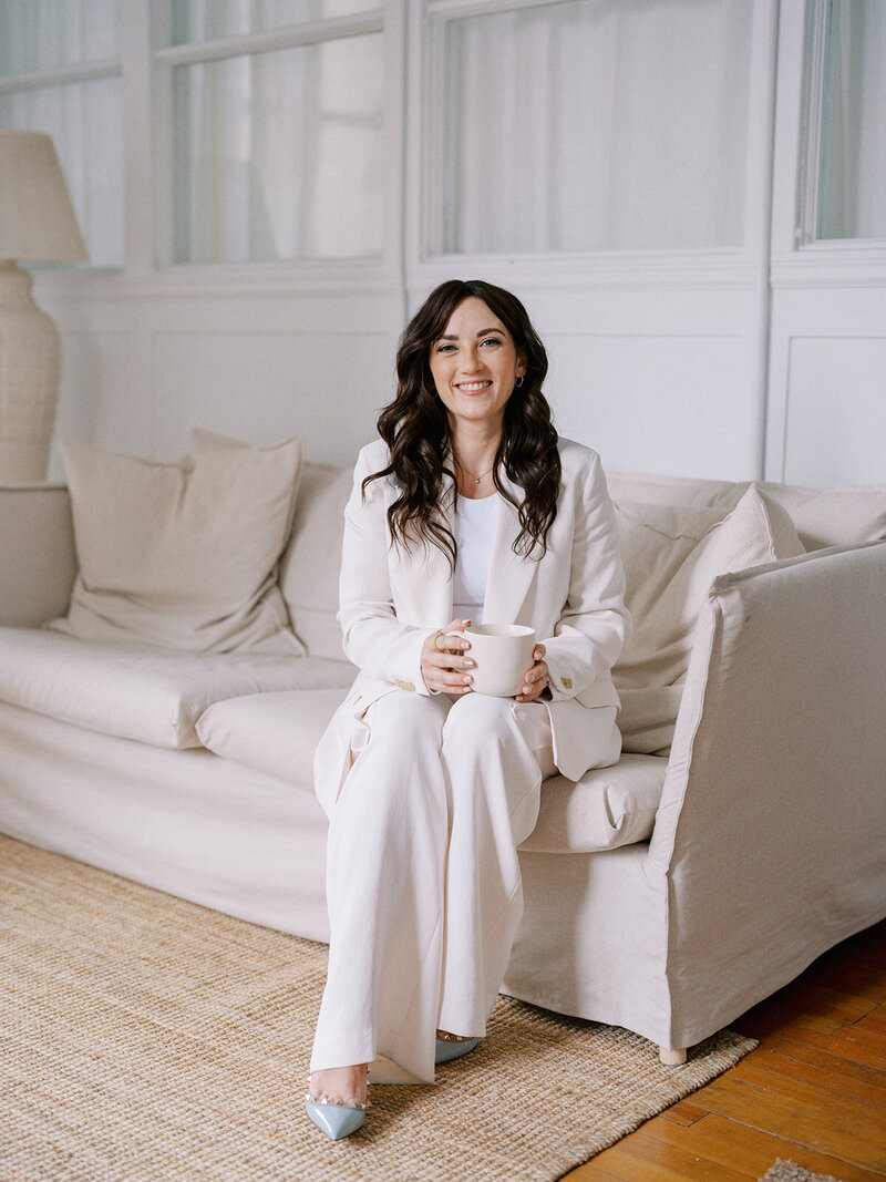 A woman with long dark hair, dressed in a white suit, sits on a beige sofa holding a mug, with a neutral expression. She is in a light-colored room with natural elements, embodying the calm precision of a Calgary wedding planner.