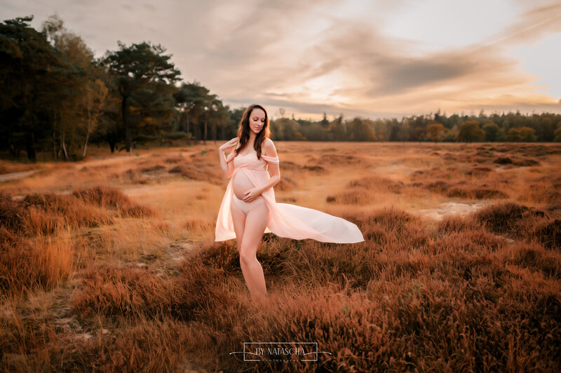 Zwangere vrouw met een roze zwangerschapsjurk aan die openvalt en haar zwangere buik zichtbaar is, kijkend langs de zijkant over haar schouder in de heide van Herperduin