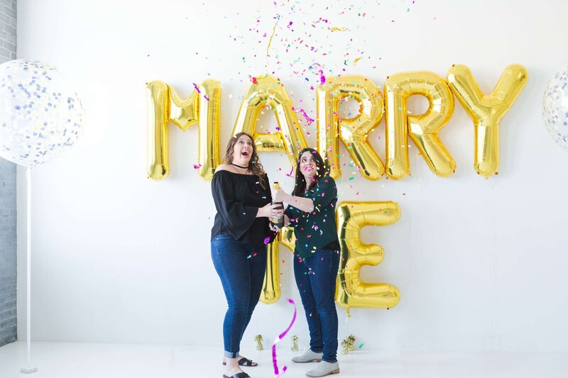 engaged lesbian couple toss confetti during their engagement session at the Houston Event Venue groom kisses bride to be on a rooftop in downtown Houston by Swish and Click Photography