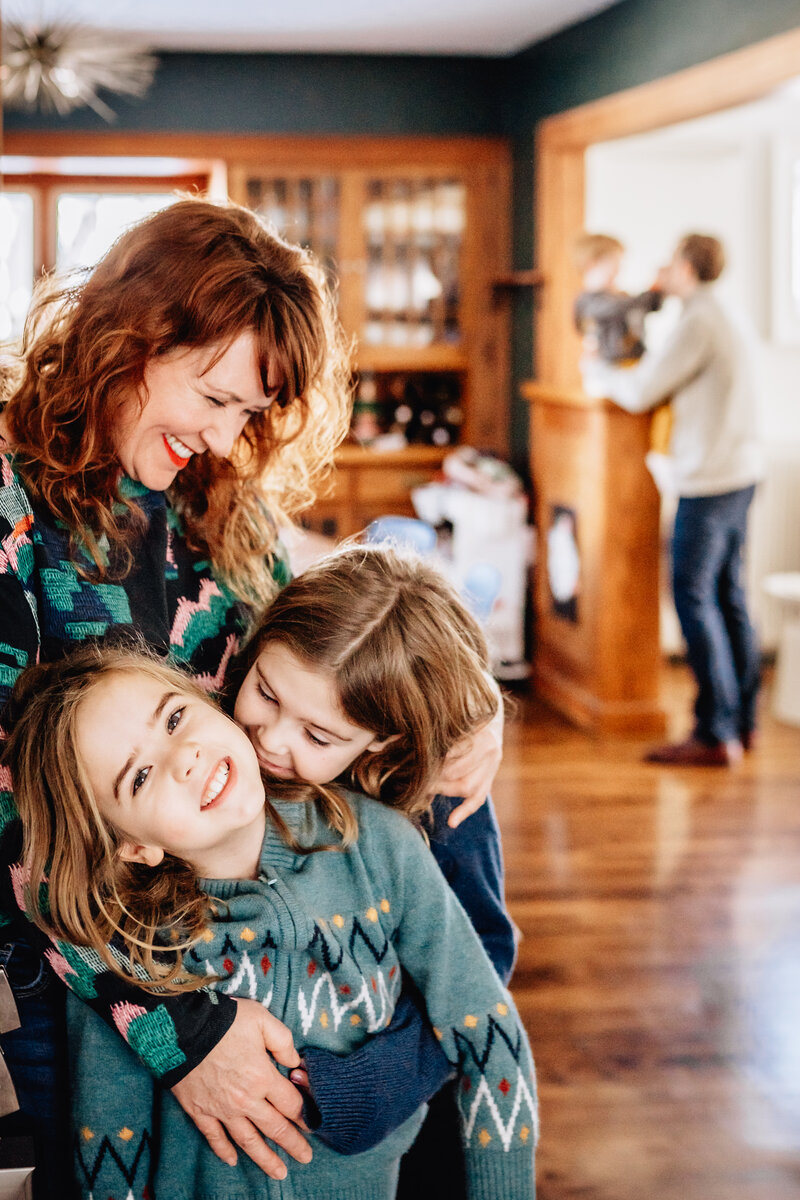 A mother joyfully embraces her two young children in a cozy, warmly lit home while another adult and child are seen in the background, creating a lively and loving atmosphere.