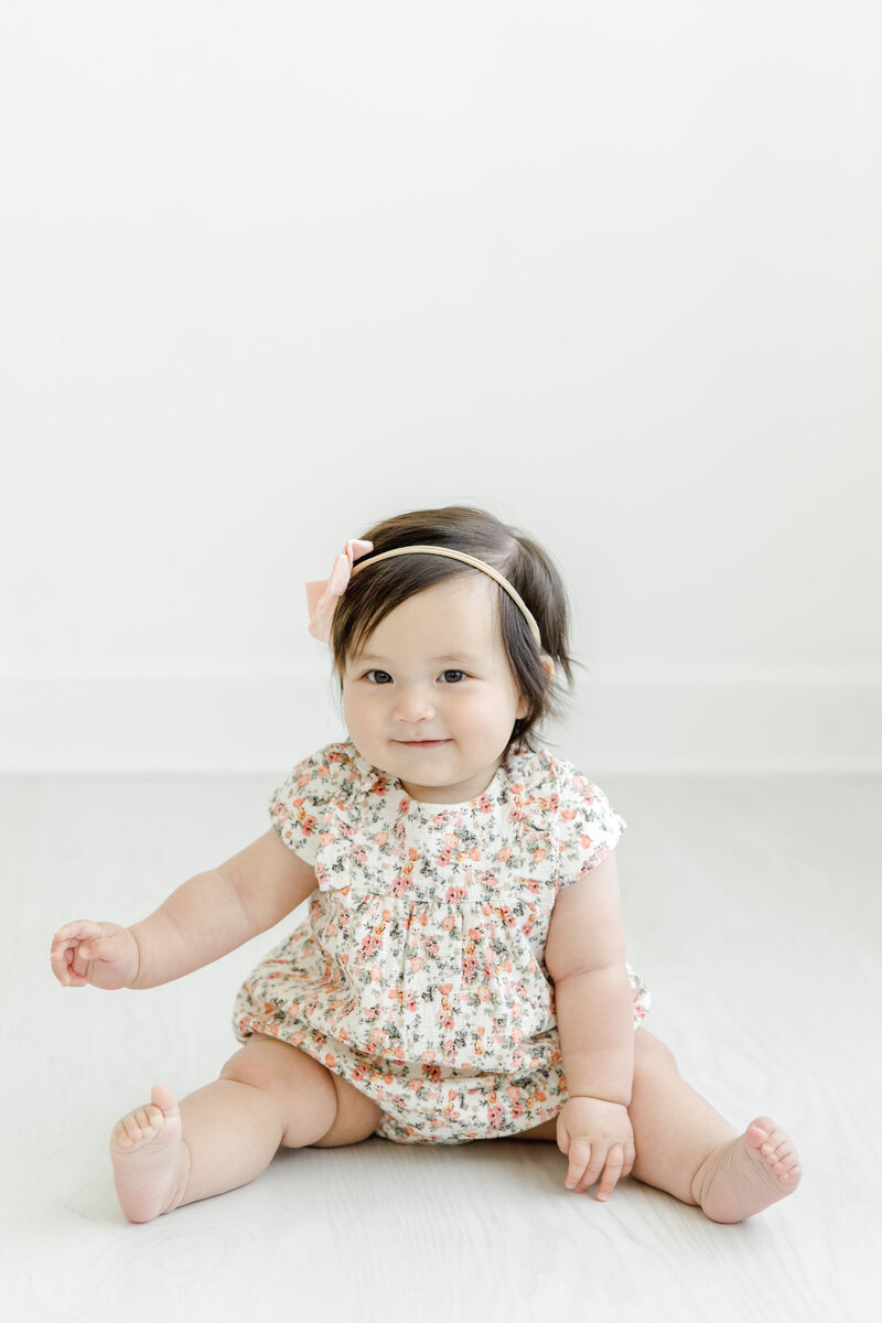Baby girl sits and smiles for portrait in a floral romper and pink headband