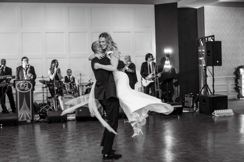 Black and white image of a groom twirling his bride off the ground. A band is playing in the background.